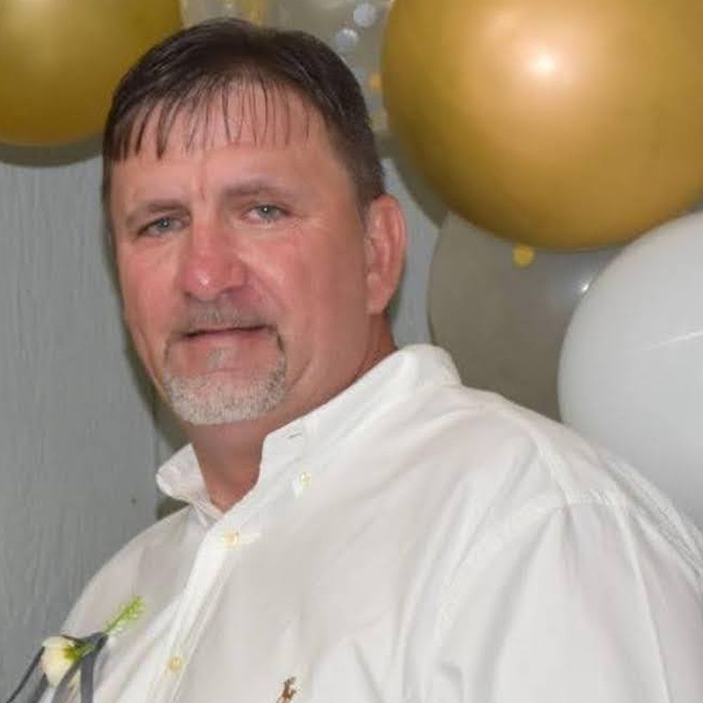 A photo shows a White man with brown hair and a gray beard. He’s wearing a white dress shirt with a boutonniere and standing in front of gold and white balloons. 