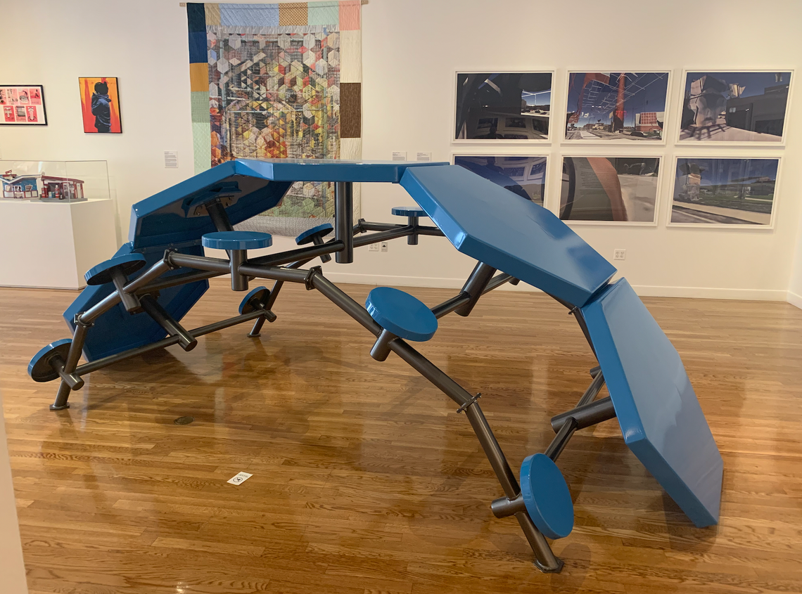 A blue and black sculpture, composed of tables and stool similar to those found in prison visiting rooms, is on display in an art exhibit. 