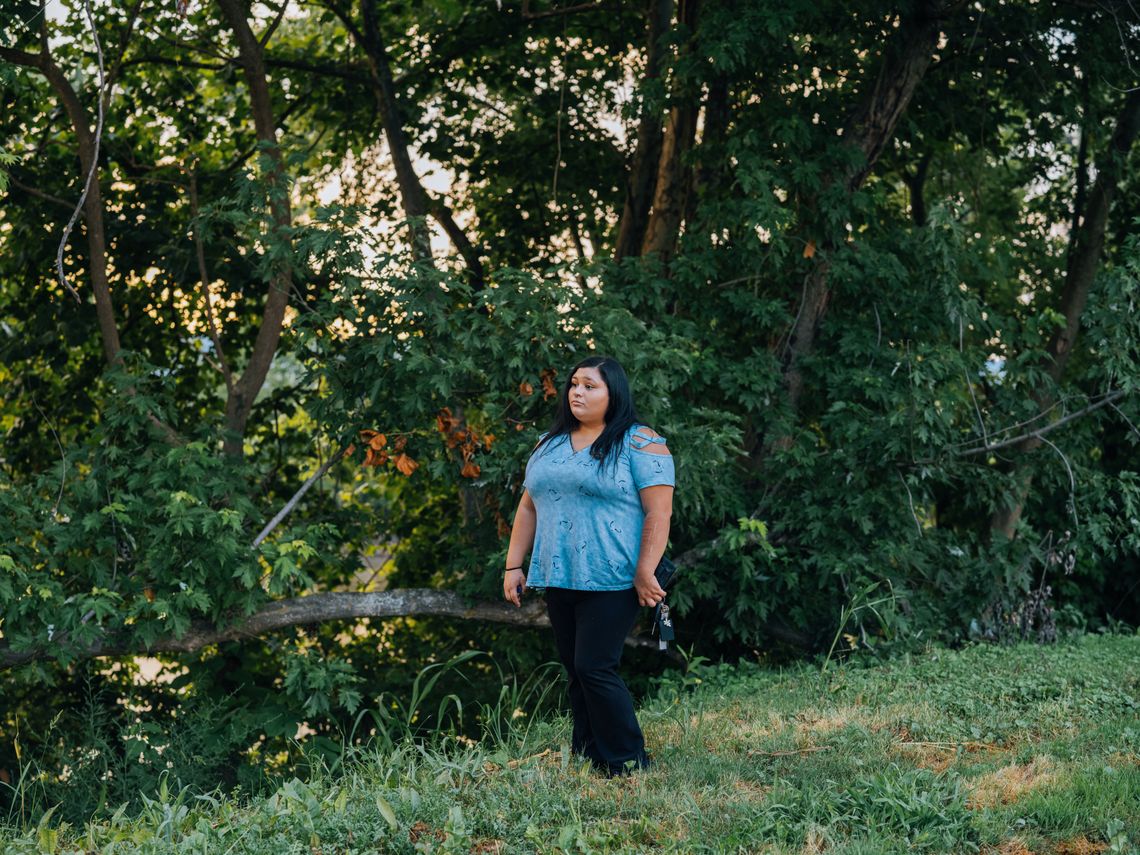A photo shows a Mexican American woman in a blue top and black pants standing in front of some leafy trees on the grass. 