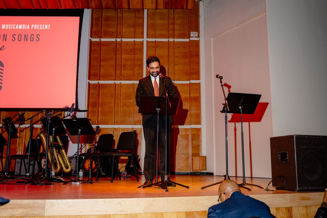 A man with dark hair and a beard in a suit with a red, dotted tie stands in front of a microphone, looking down at a lectern. He stands on a stage and behind him is visible wooden acoustical tiles hanging on the wall and a number of music stands, chairs and saxophones meant for musicians to sit in and perform. In the foreground, a bald man with glasses is visible, sitting in the audience in front of the stage.
