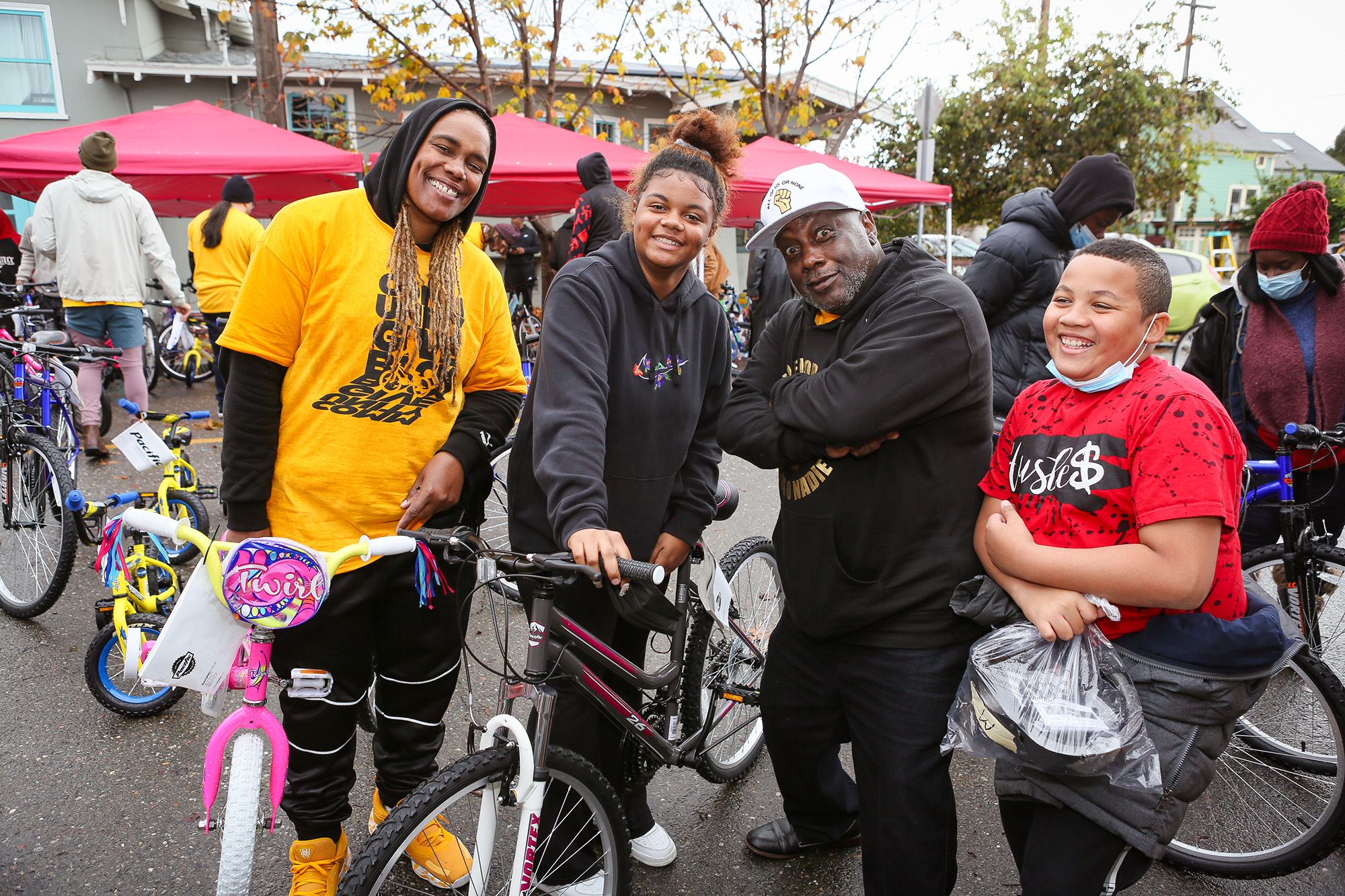 Four Black people smile as they pose for a picture while standing next to bikes. Community members look at several bikes in the background. 