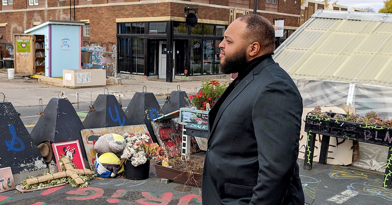 A Black man, wearing a black suit, looks to the left while standing in front of a memorial on a street. 