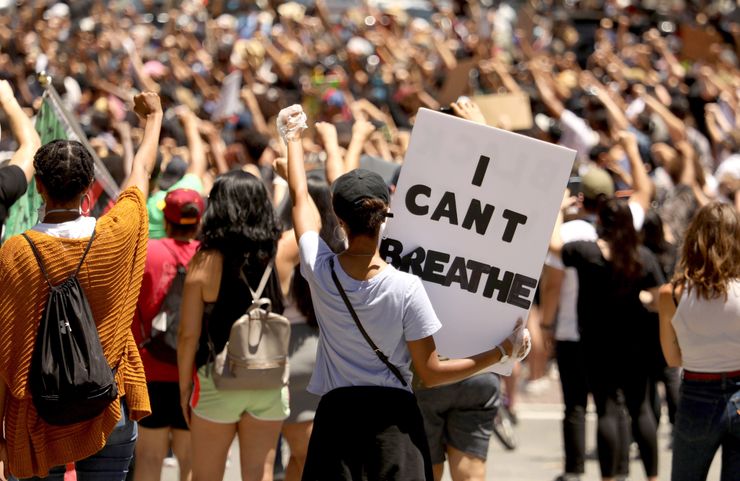 A woman with medium-dark skin tone stands during a protest, raising her fist and holding a sign that says, "I can't breathe." Hundreds of individuals also are raising their fists in the background. 

