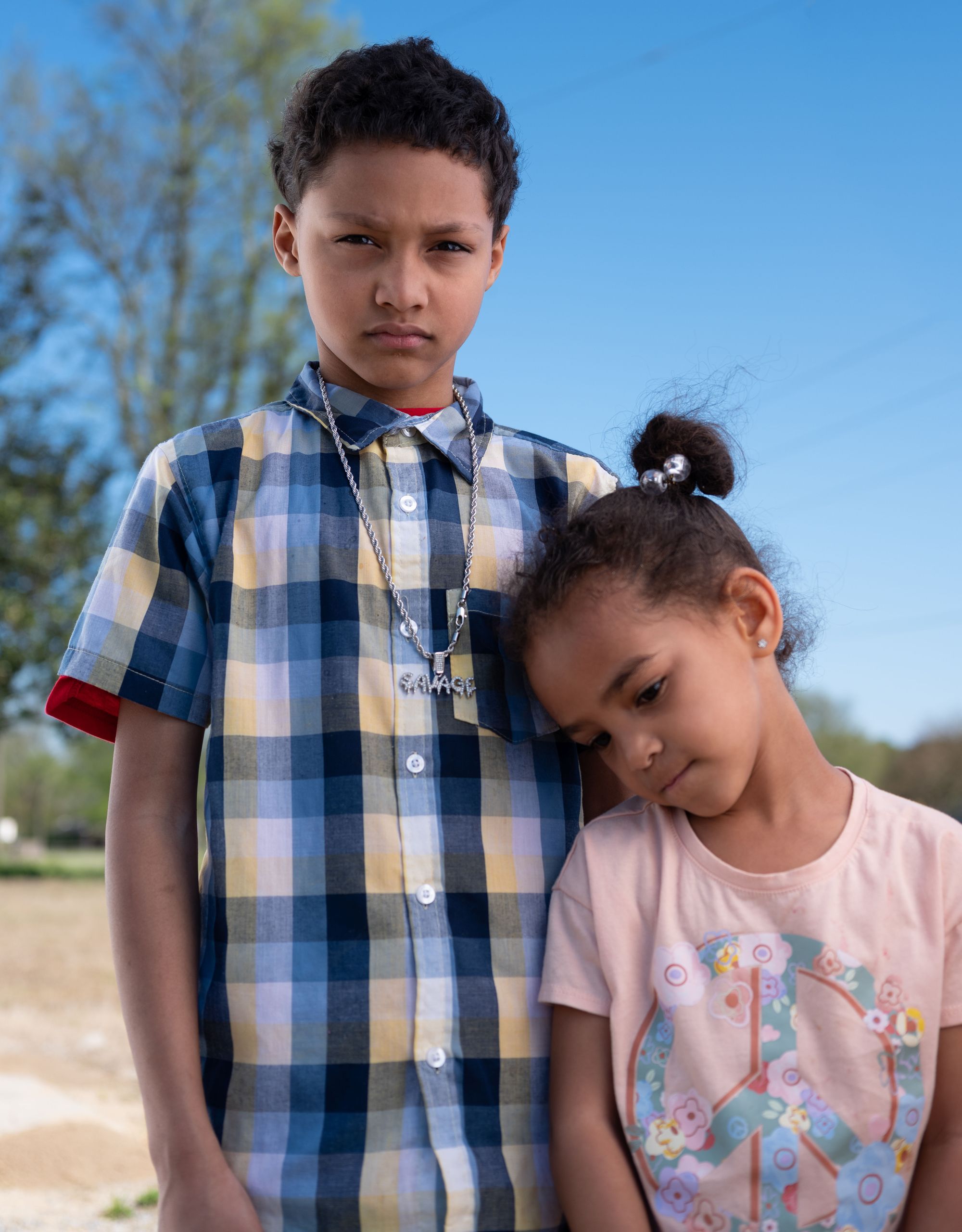 A young multiracial boy wearing a blue and yellow plaid shirt, stands next to his cousin, a young multiracial girl wearing a pink T-shirt, who is leaning her head on his chest. 