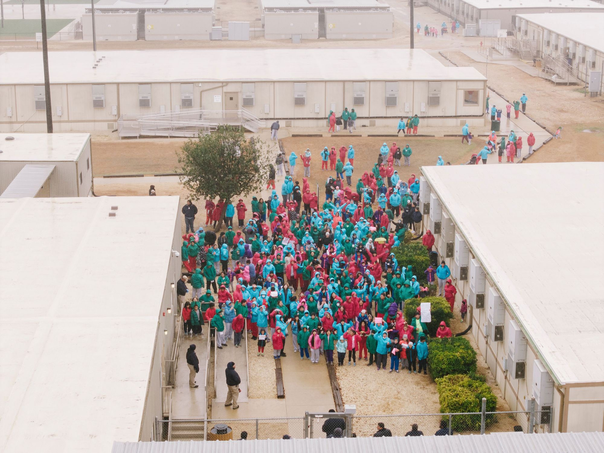 A photo shows a crowd of adults and children in turquoise, red, pink and green coats standing between buildings on a detention center campus. Some people are holding up signs. 