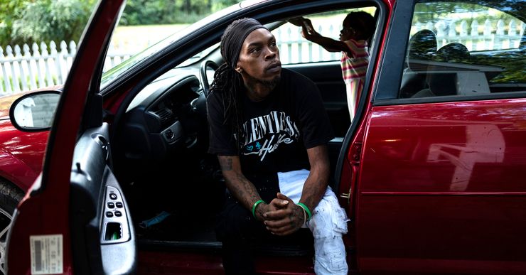 A Black man, wearing a black headband, black graphic T-shirt and black and white jeans, sits in the driver’s seat of a red car with the door opened. His young daughter plays inside the car in the background. 