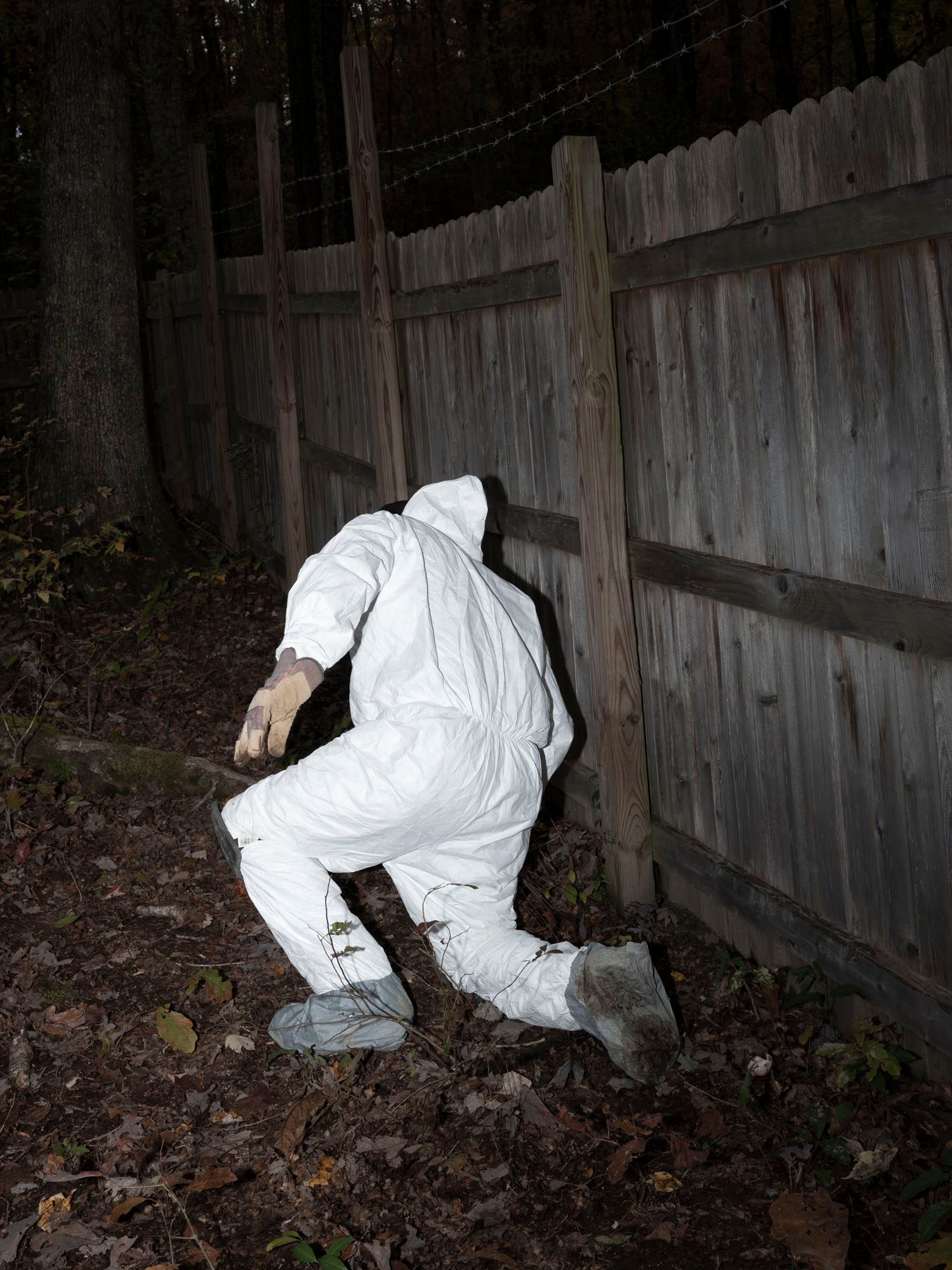 A student dressed in a white hazmat suit kneels on the ground near a fence outdoors. 