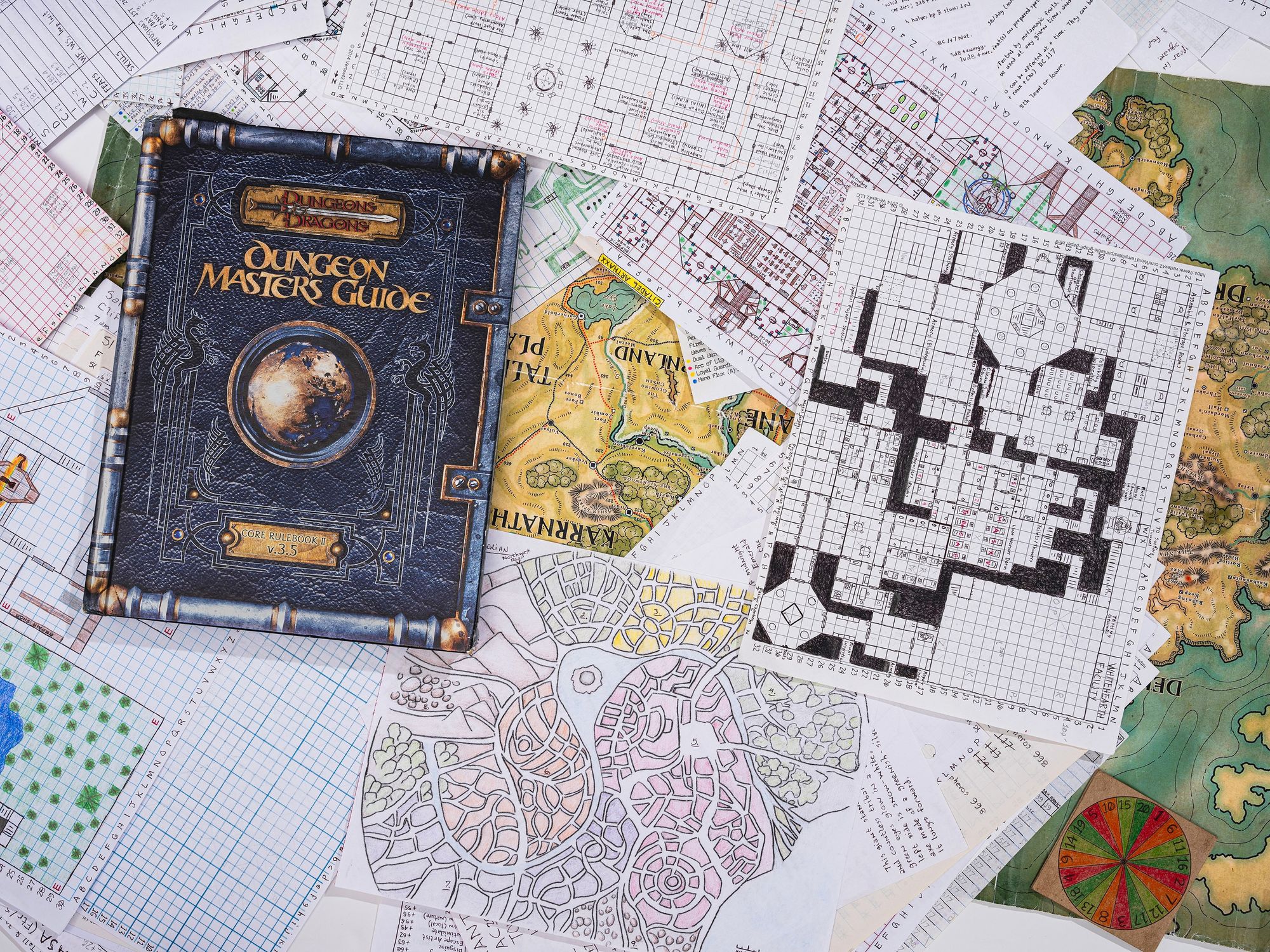 A Dungeons and Dragons book titled “Dungeon Master’s Guide” sits atop a pile of scattered maps and drawings. Many of the hand-drawn maps are sketched on graph paper. Some pages contain details in color, and others are drawn in black-and-white. 