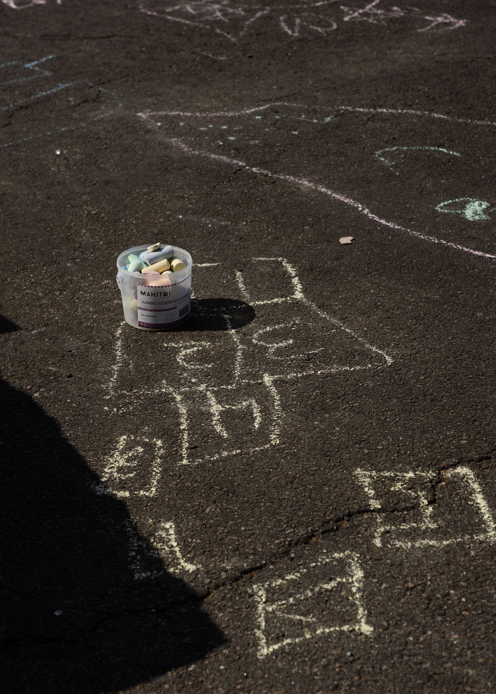 A photo shows chalk drawings of hopscotch squares on asphalt. A tub of chalk is visible in the middle of the photo. 