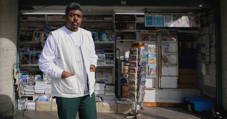 Donovan X. Ramsey, a Black man with short hair, wears a white shirt, vest and green pants, and stands in front of a newsstand.  