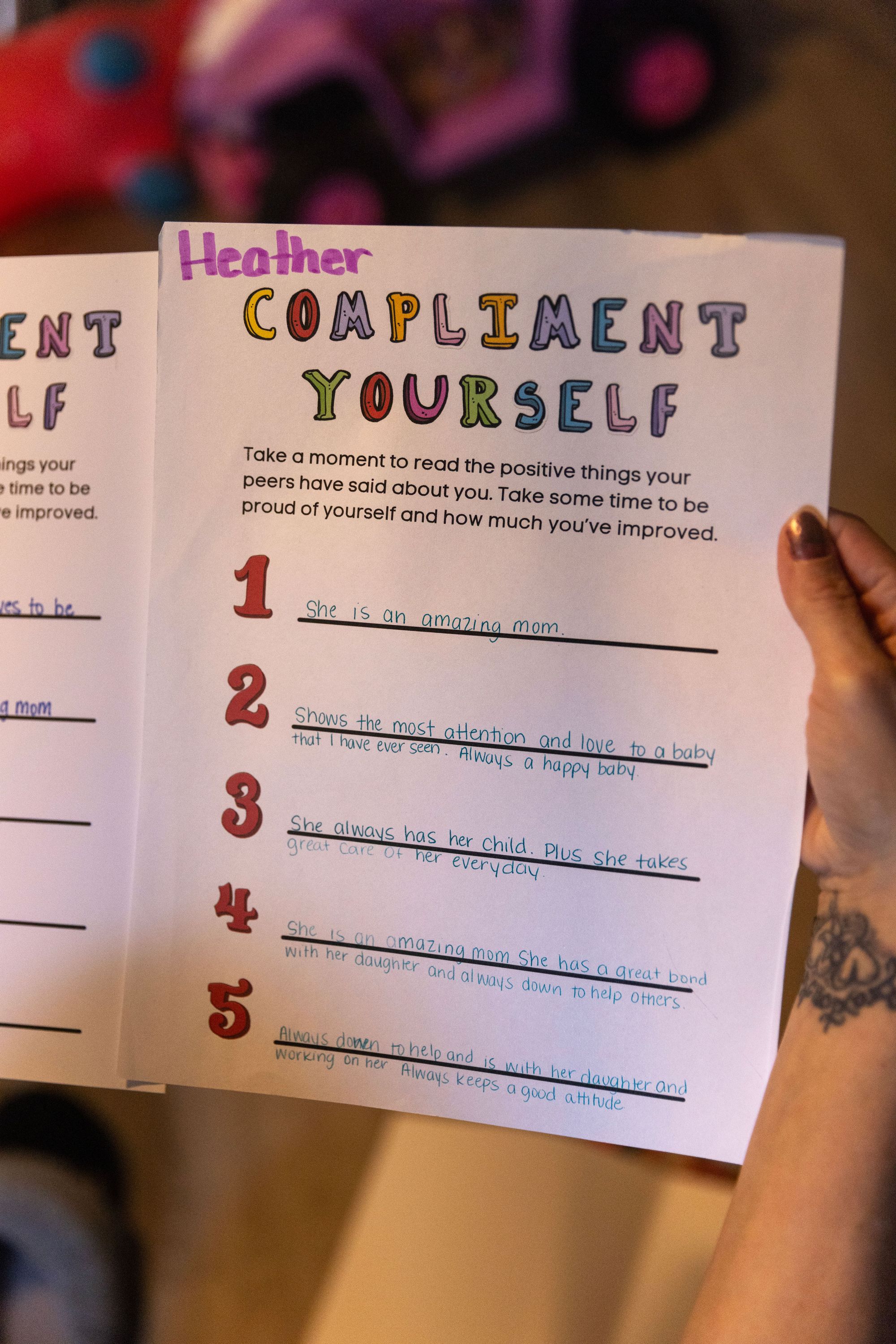 A close-up photo shows a White woman’s hand holding a worksheet that reads “COMPLIMENT YOURSELF” at the top. The name “Heather” is handwritten in the top left corner of the page. The worksheet shows a list of five handwritten compliments, like, “She is an amazing mom.”