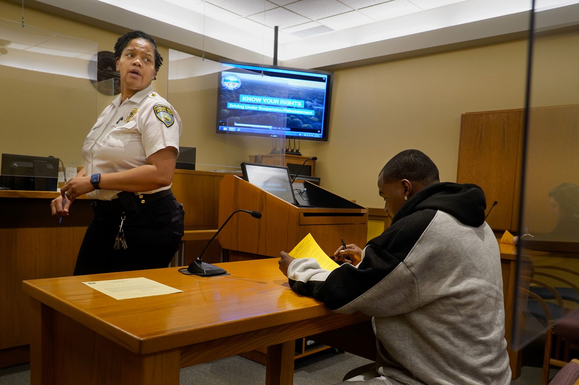 A Black woman in a bailiff’s uniform looks over her shoulder while walking, as a Black man in a black and gray hoodie sits at a desk to fill out a form.