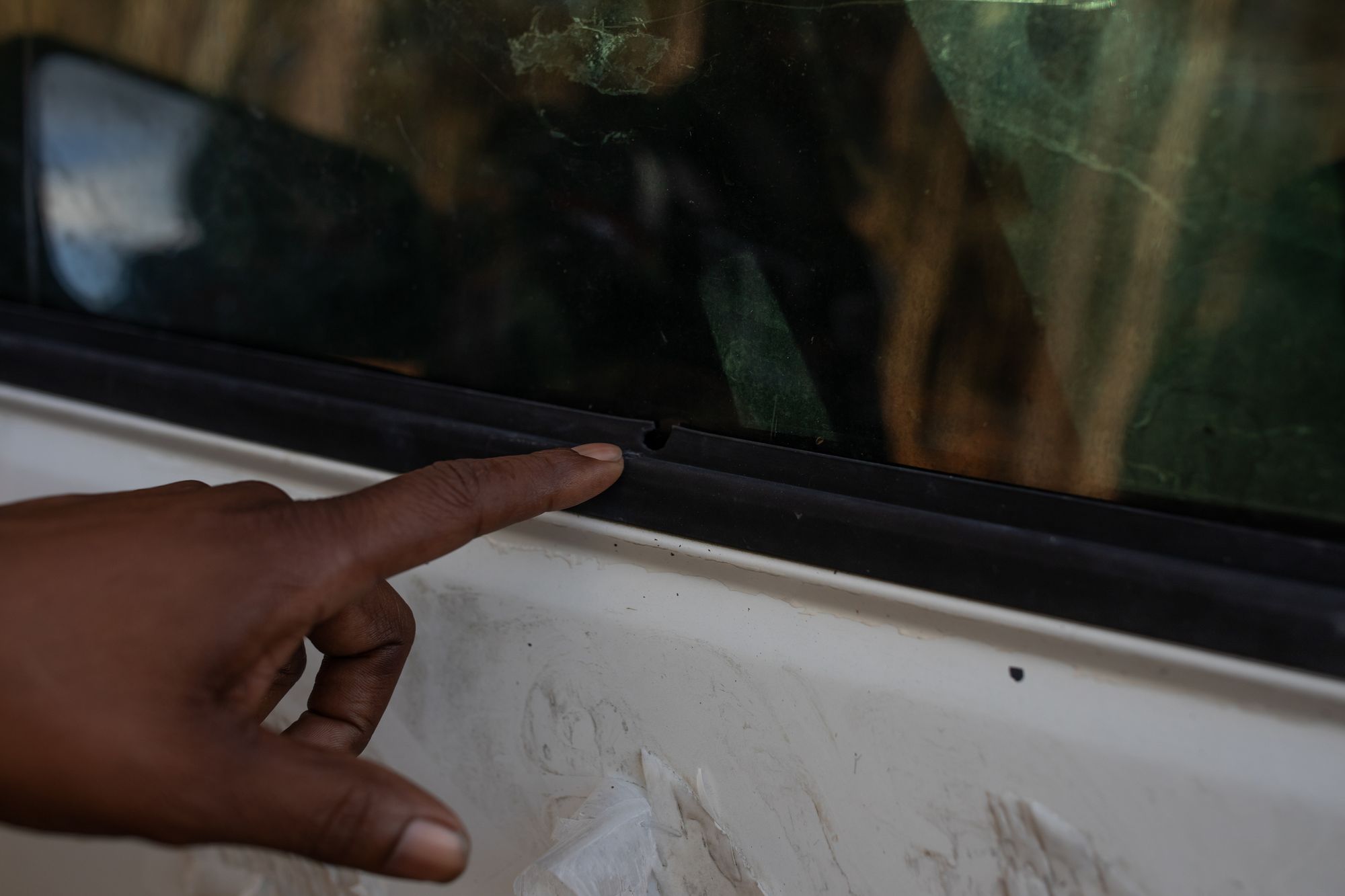 A photo shows the finger of a Black man pointing at a bullet hole in the weather stripping of a truck’s window. 