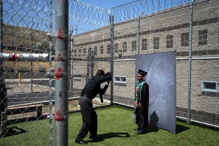 A photographer takes a picture of an incarcerated man, wearing a graduation cap, gown, a Mexican sarape print stole and a green stole that reads, "T.O.P.S.S." in front of a gray backdrop. The picture was taken outside on a lawn surrounded by barbed wire fences.