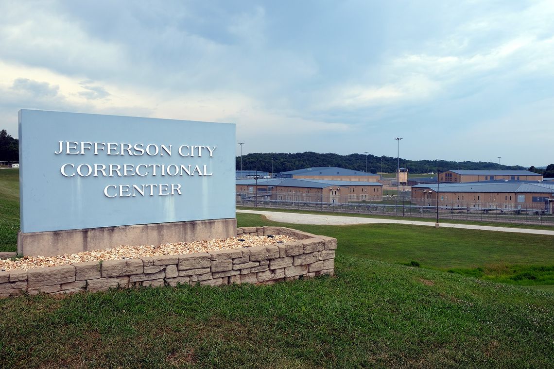 An image shows a light blue sign that reads "Jefferson City Correctional Center." Gray and tan prison buildings surrounded by wired fences are in the background. 