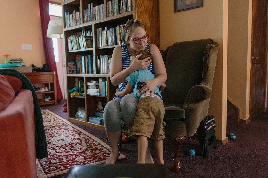 Grace Smith, a White woman wearing glasses, a blue and white striped tank top and gray leggings, hugs her child Julian while sitting on a sofa chair. 