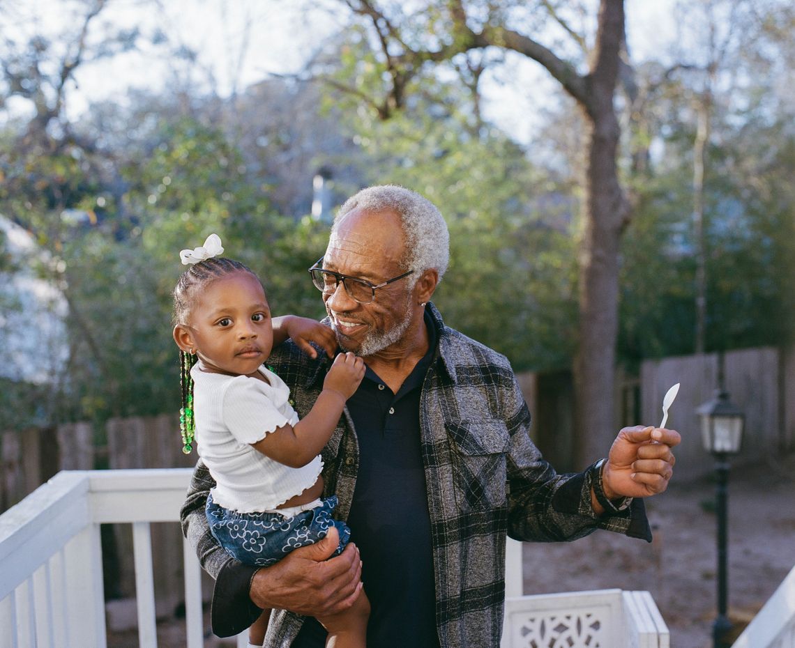 Moses stands on a deck, smiling at and carrying Armour’s great-niece, a Black toddler wearing a small white bow and braids in her hair.
