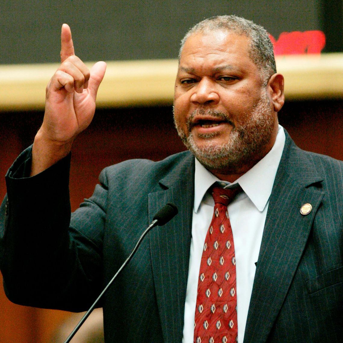 A color photo of a Black man wearing a suit. The man is speaking into a microphone and pointing upward. 