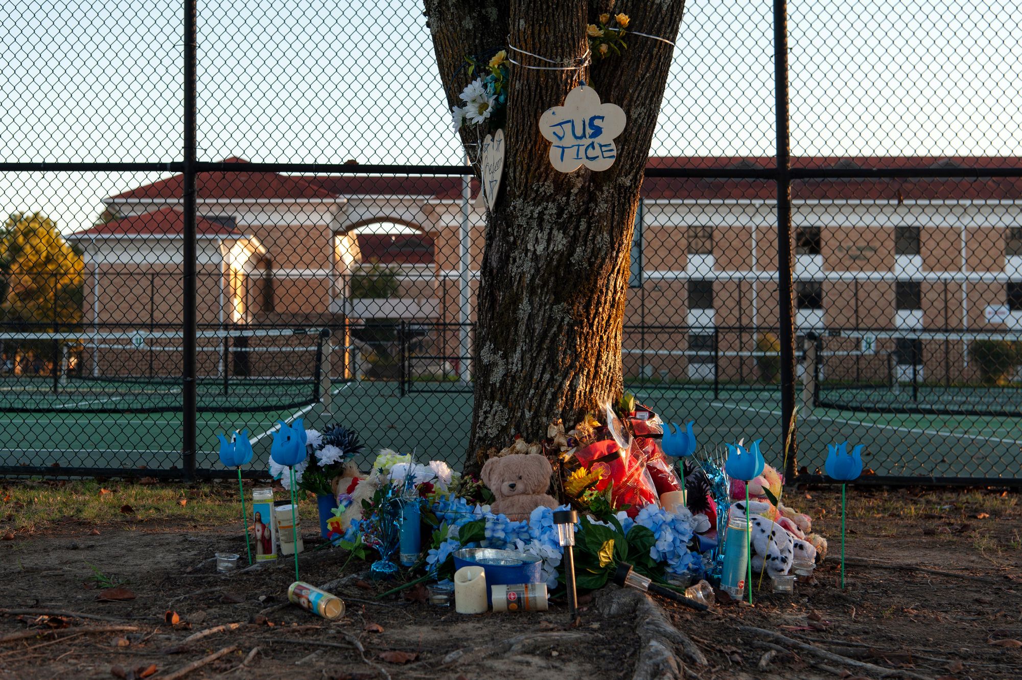 A photo shows a tree in front of a college athletic field and building. Blue flowers, candles and memorial notes for Trey Reed surround the bottom of the tree. 