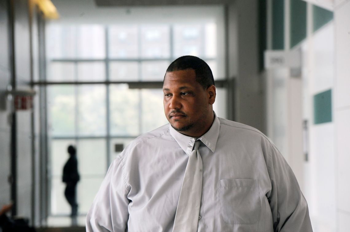 A Black man wearing a light grey shirt stands in the Bronx Criminal Court building. 