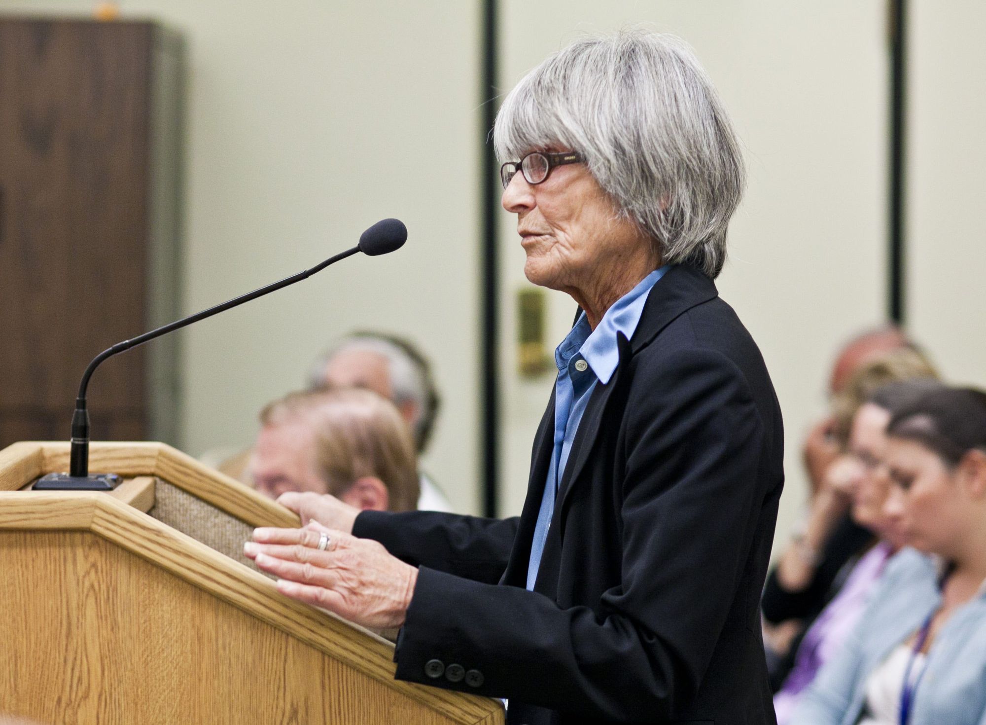 Judy Goldman, a White woman, spoke at a podium to a group of people in a meeting room.