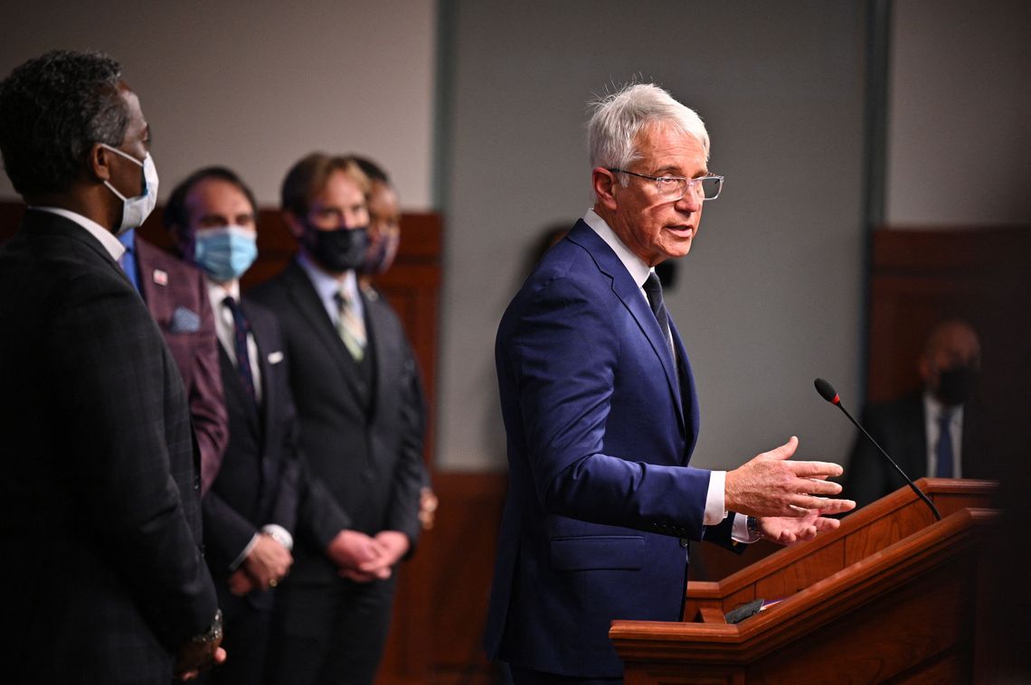 Los Angeles District Attorney George Gascón, with white hair and glasses, stands at a podium on the right, wearing a blue suit and white shirt. A group of people stand behind him at the left.