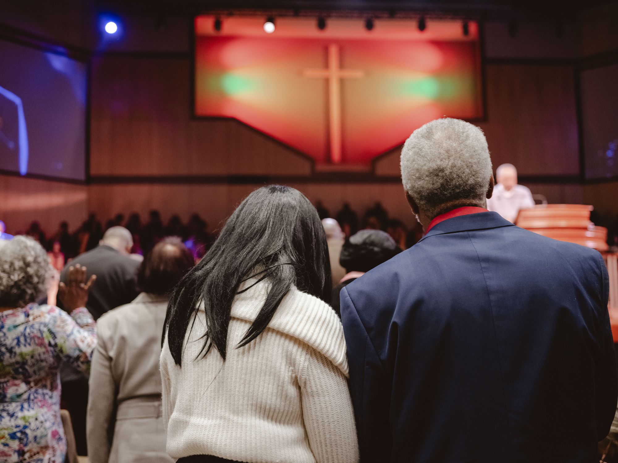 The backs of Armour and Moses are visible, as they look at the podium in a church. In the background is a banner with a cross on it. 