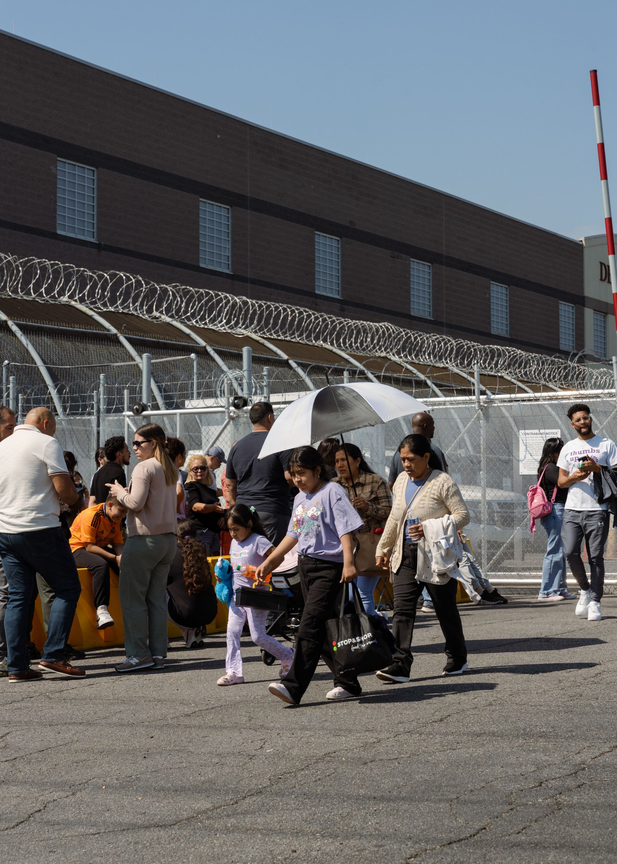 A photo shows people walking and standing outside Delaney Hall, in front of barbed wire fencing. Behind the barbed wire fencing is a building with square windows. 