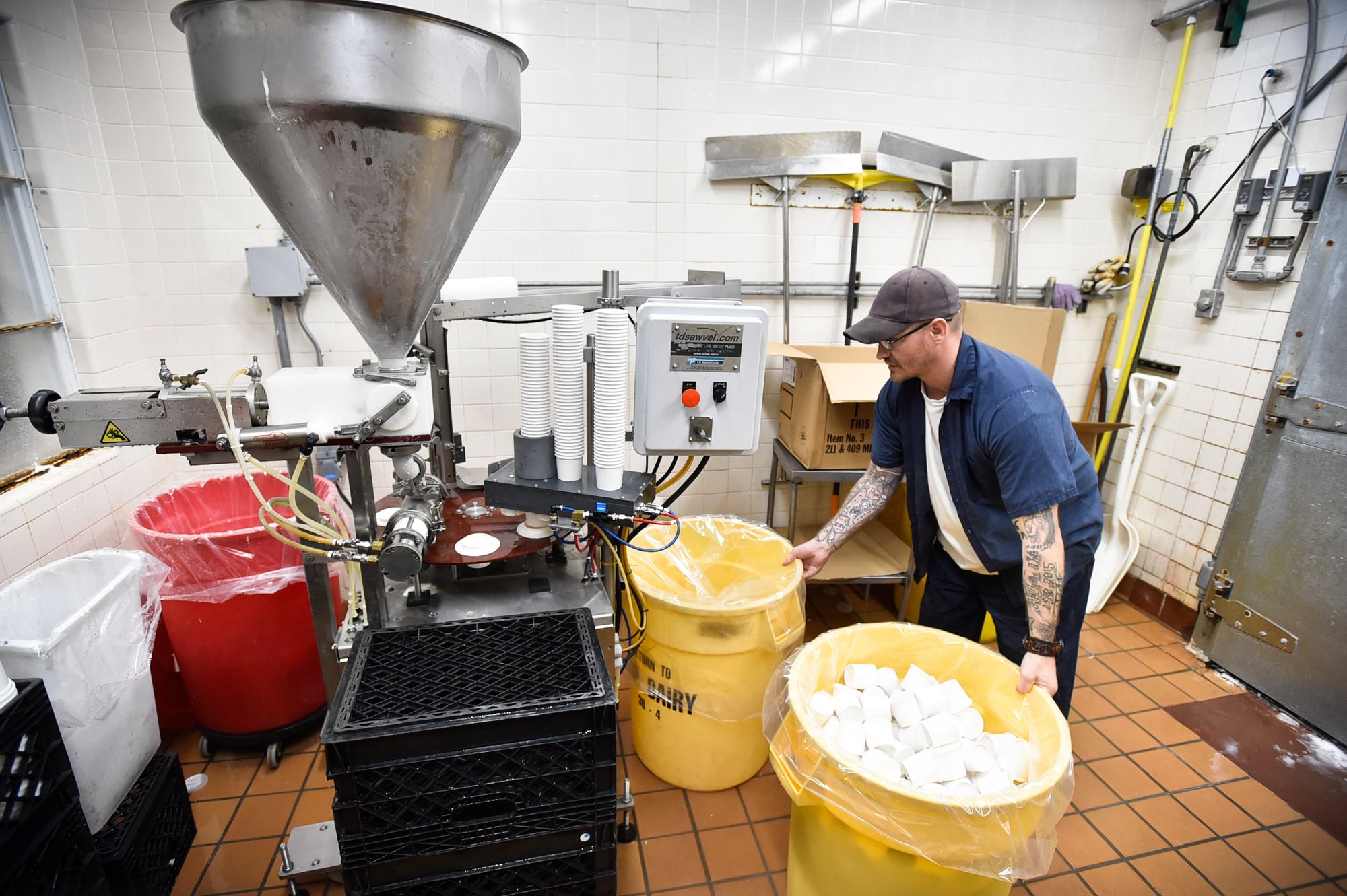 A man, wearing a blue prison uniform, white T-shirt, tan cap and glasses, holds onto two yellow bins in front of an industrial ice cream machine. 