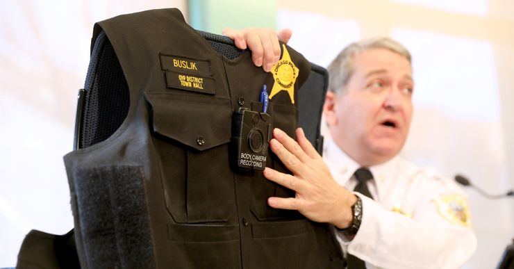 A White male police officer holds a police vest, pointing out its body camera.  