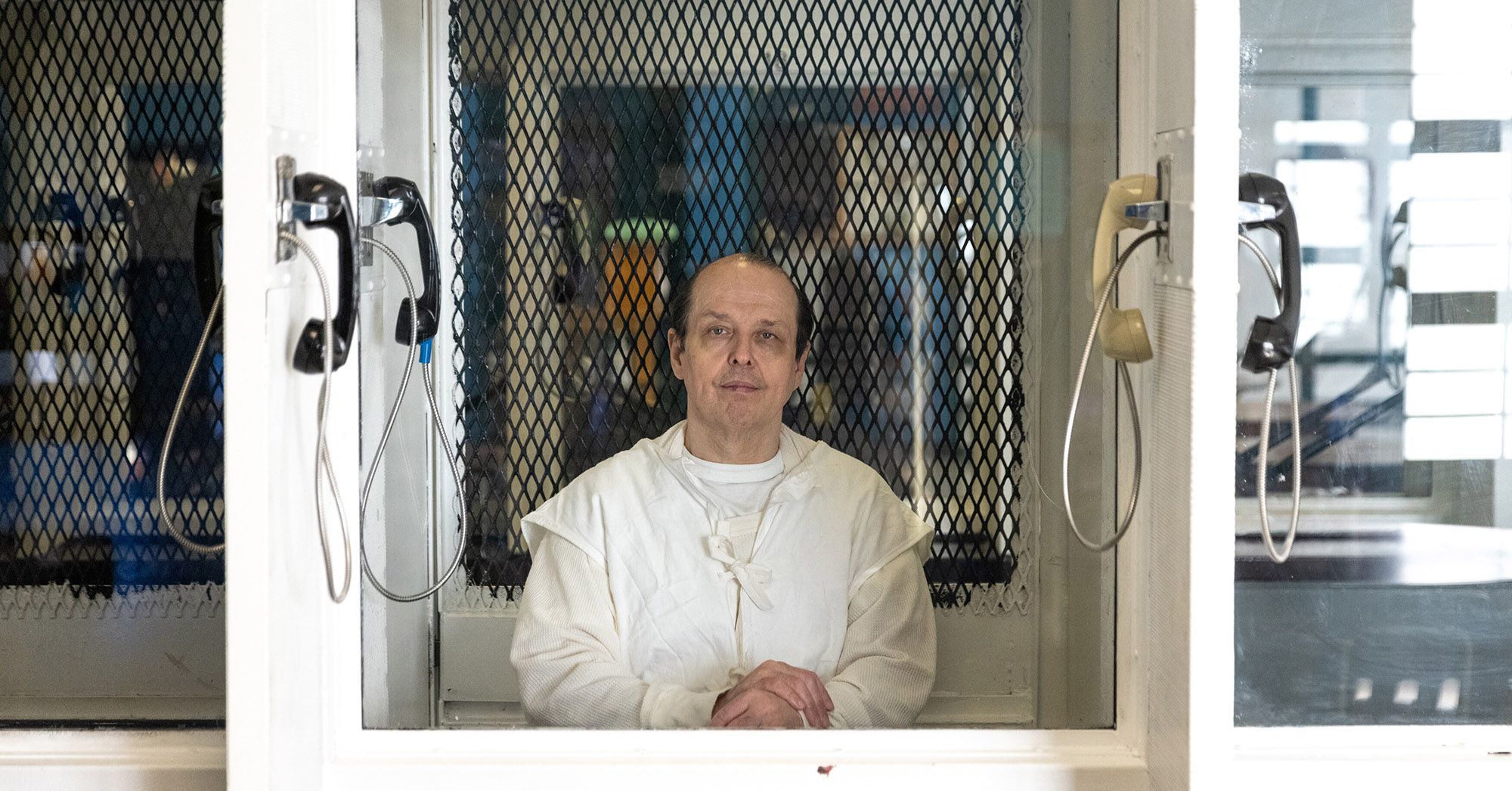 Robert Roberson, a light-skinned man wearing a white prison uniform, looks through visitation room glass. A phone hangs on either side of the divider.