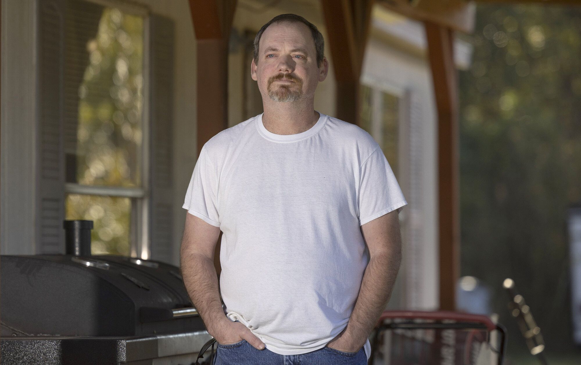  A White man with a goatee, white T-shirt and short, dark hair stands in front of a barbecue grill with his hands in the front pockets of his blue jeans.
