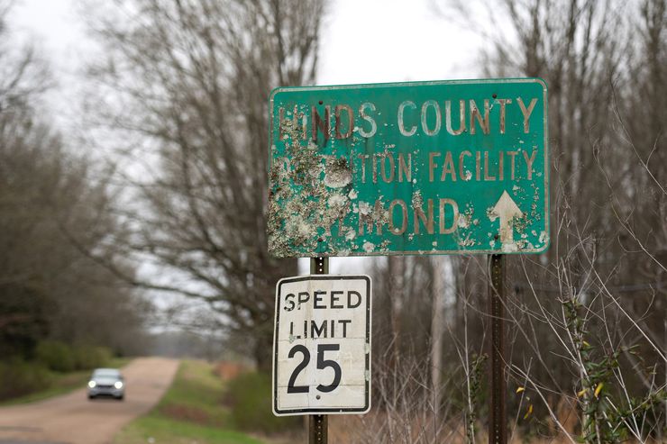 A photo of a damaged green road sign that reads “Hinds County Detention Facility Raymond” with an arrow pointing forward. 