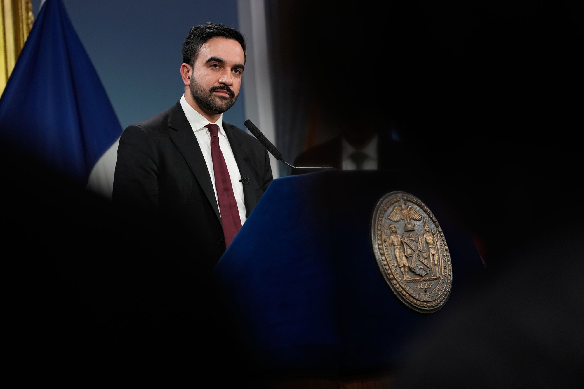 A photo of Zohran Mamdani, a light-skinned man with black hair and beard in a dark blue suit, maroon necktie and a white dress shirt, standing at the podium at a news conference with dark silhouettes framing the picture in the foreground. 