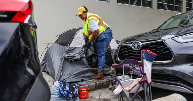 A dark-skinned woman, wearing a reflective safety vest, white T-shirt, blue jeans, yellow cap and gloves, takes items out of a tent located on a sidewalk. A camping chair, a backpack, a plastic bag and a water jug are in front of the tent. Two cars are parked next to the tent. 