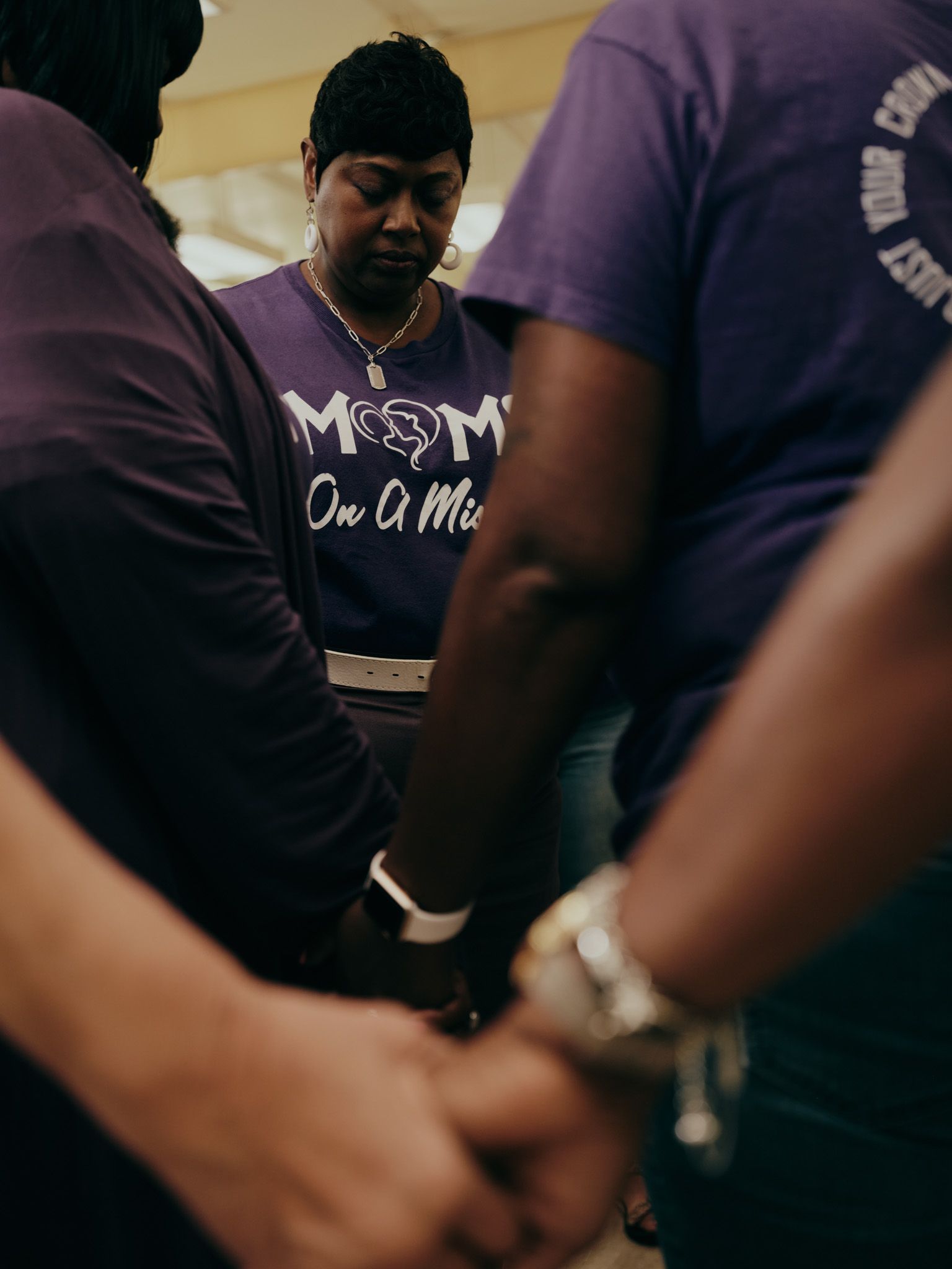 A vertical photo shows people in a circle of healing, holding hands. All are wearing purple T-shirts that say “Moms on a Mission.” Lennie Ross, a Black woman with short-cropped hair situated in the center of the circle, tilts her head downward and has her eyes closed.