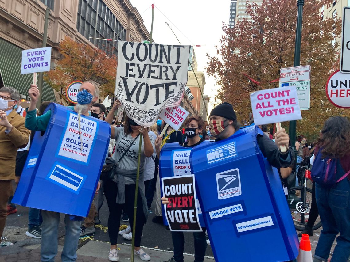 A photo shows three protesters in blue U.S. Postal Service mailbox costumes, one of which has "Mail-in ballot drop off" printed on it. They are masked. Some of the protesters hold signs saying "Every Vote Counts," "Count all the votes." 