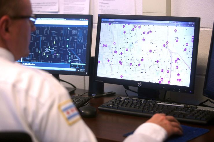 A person with light skin tone, wearing a white uniform with a Chicago flag on their shoulder, looks at gunshot detection programs on two computer screens.