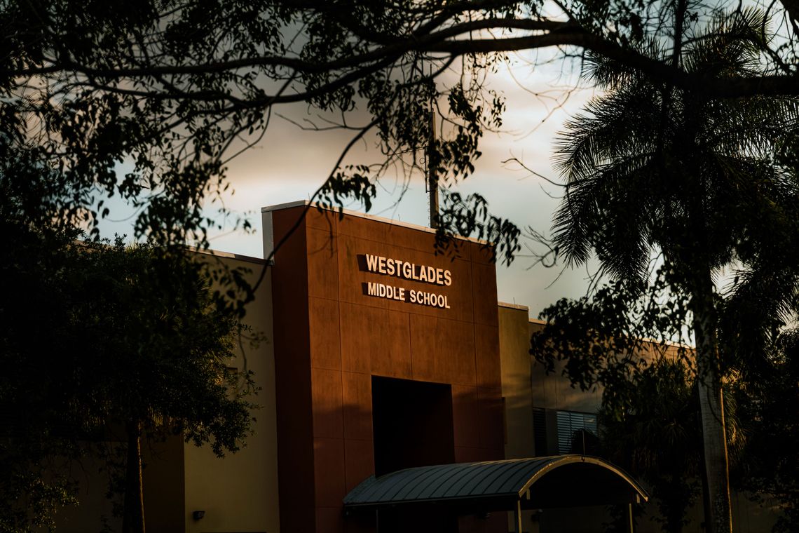 A photo of a building displaying the words “Westglades Middle School” seen through the silhouette of trees in golden afternoon light. 