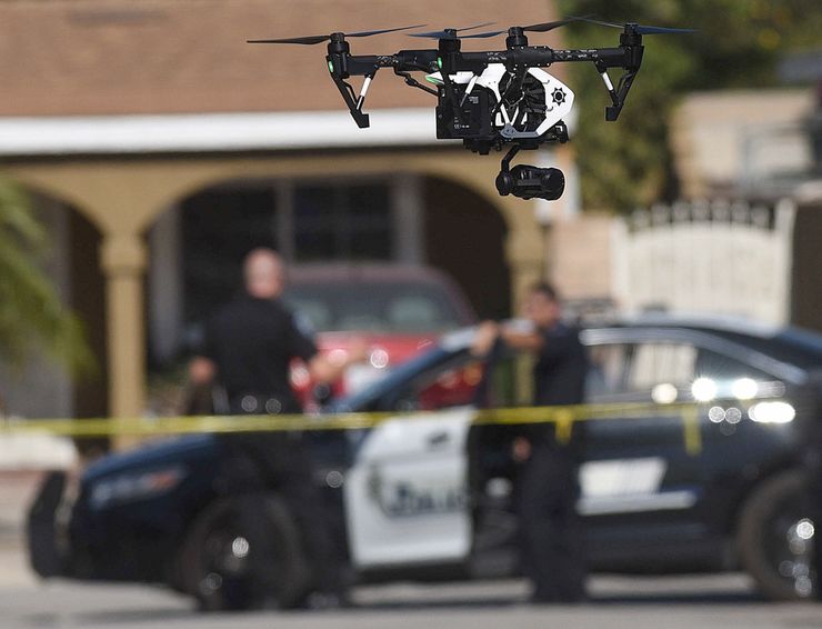A black and white drone, with a police emblem on it, flies around a crime scene. The background is out of focus, with officers standing behind yellow tape and by a parked police car. 
