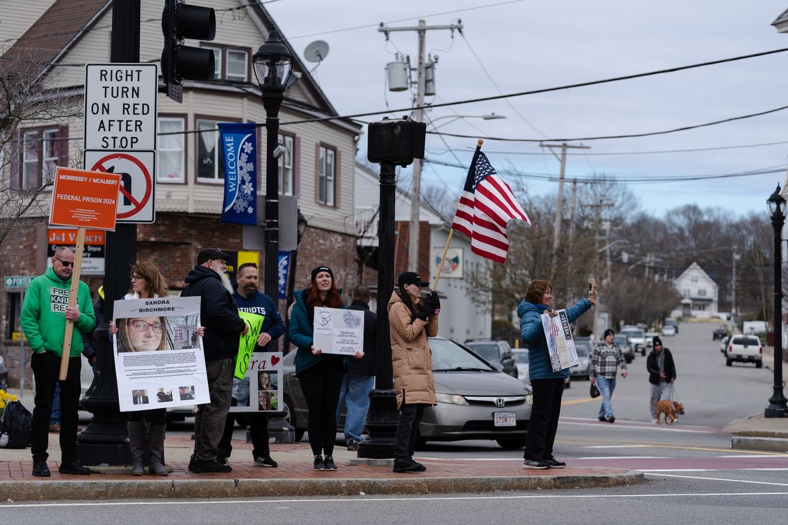 Seven people stand with signs or an American flag at a street corner in Stoughton, Mass. Some of the signs show photos of Sandra Birchmore.