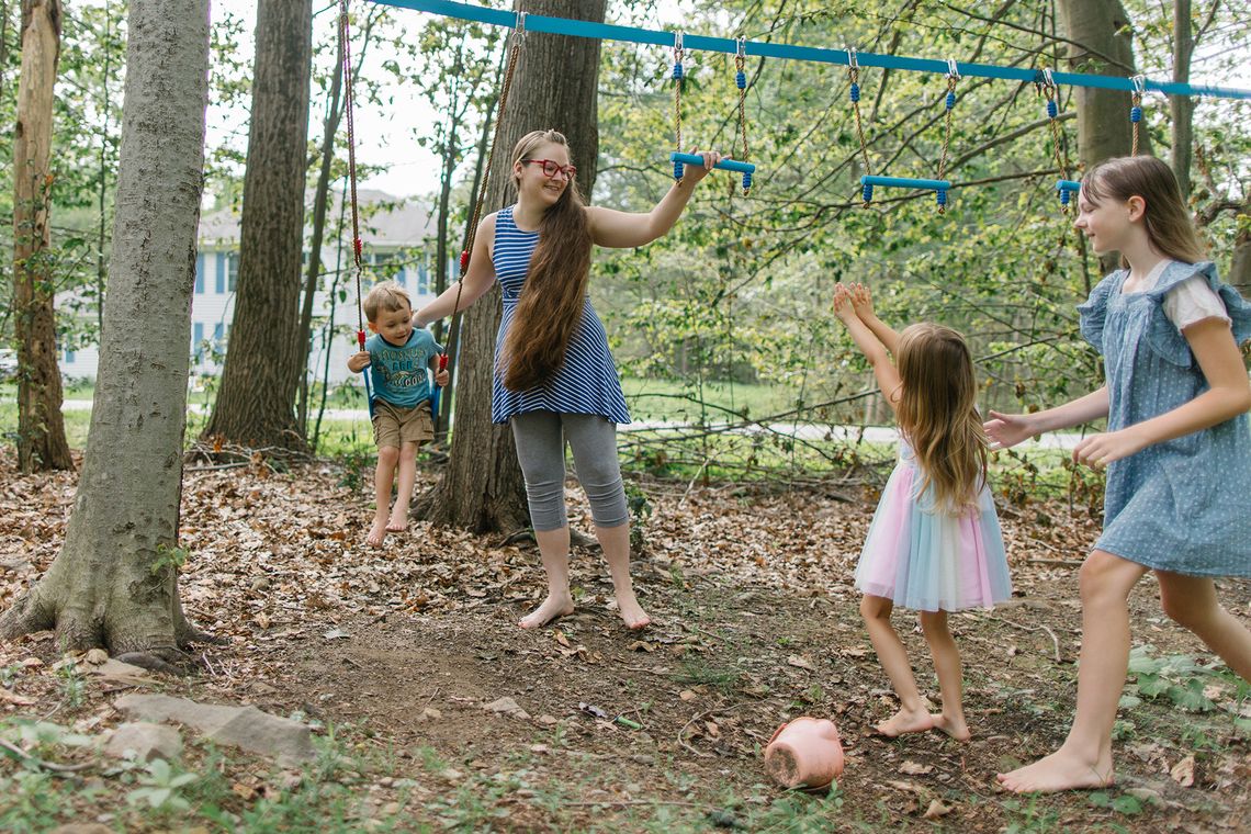 Grace Smith, a White woman wearing glasses, a blue and white striped tank top and gray leggings, pushes one child on a swing while talking to her other children. 