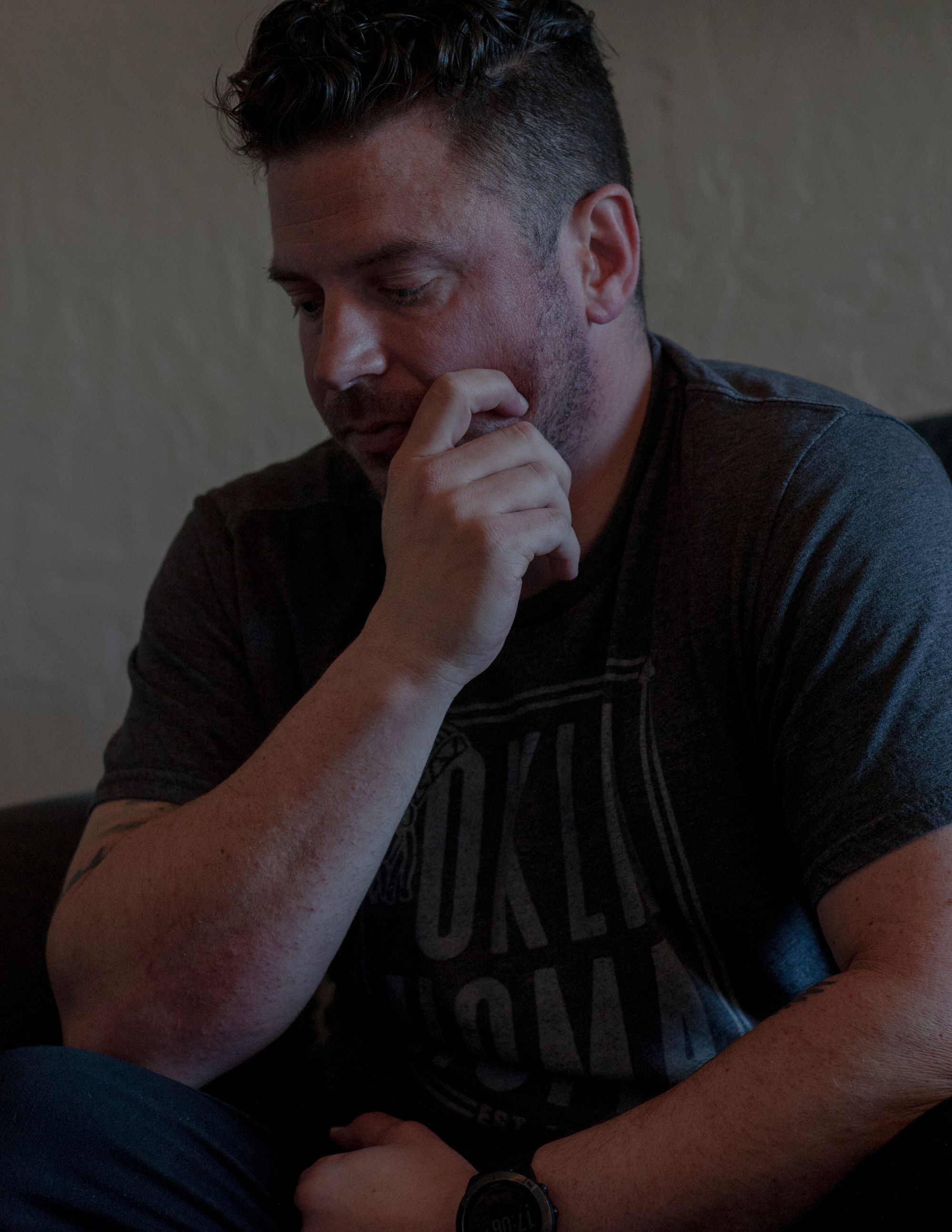 A White man in a dark gray T-shirt is sitting, looking down and to his right, while his right hand grips his jawline. 