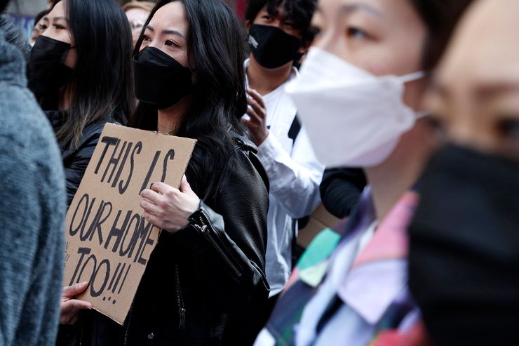 An Asian woman, standing in a crowd, holds a sign reading "This is our home too!!!" 