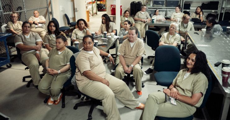 A group of women in tan prison uniforms sitting in a room, watching a television show.