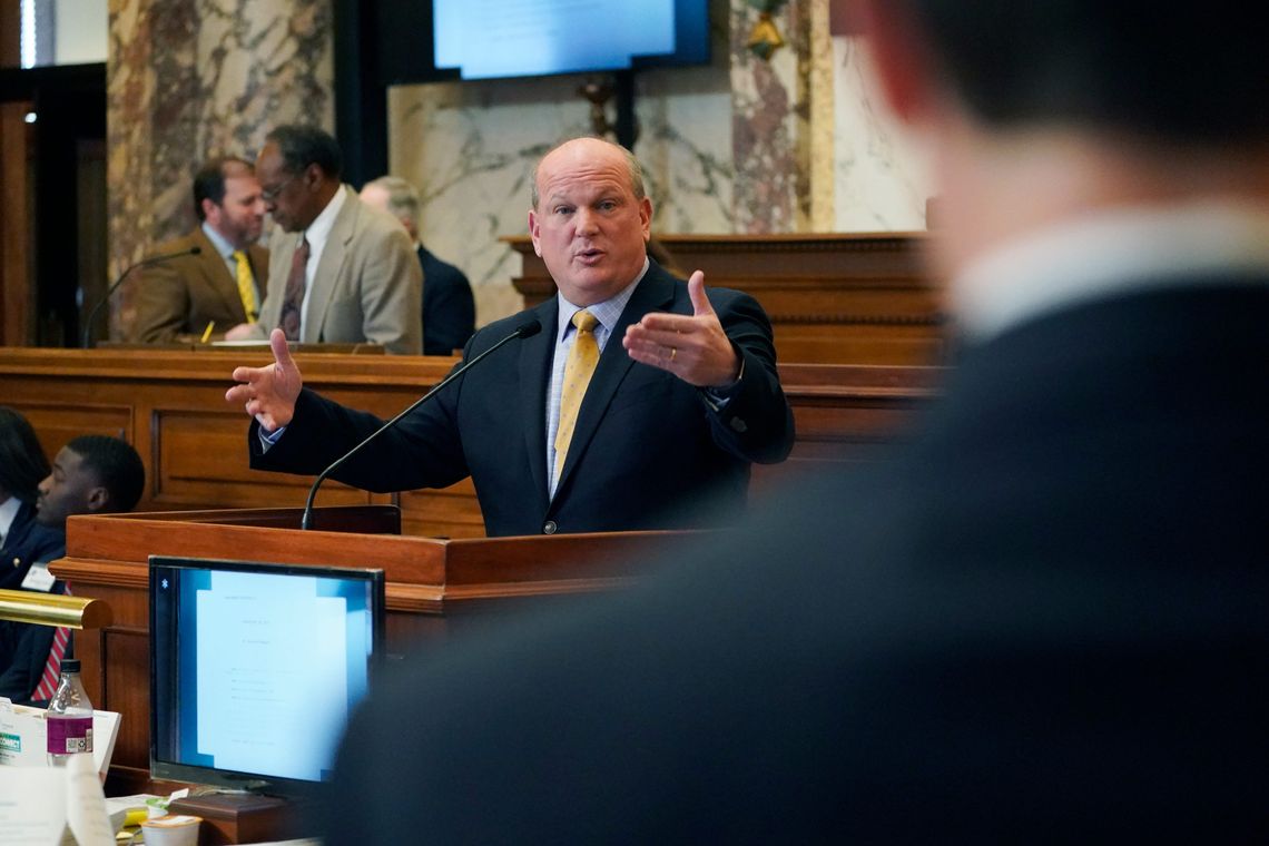 A photo of a White man in a suit gesturing toward someone while speaking. 