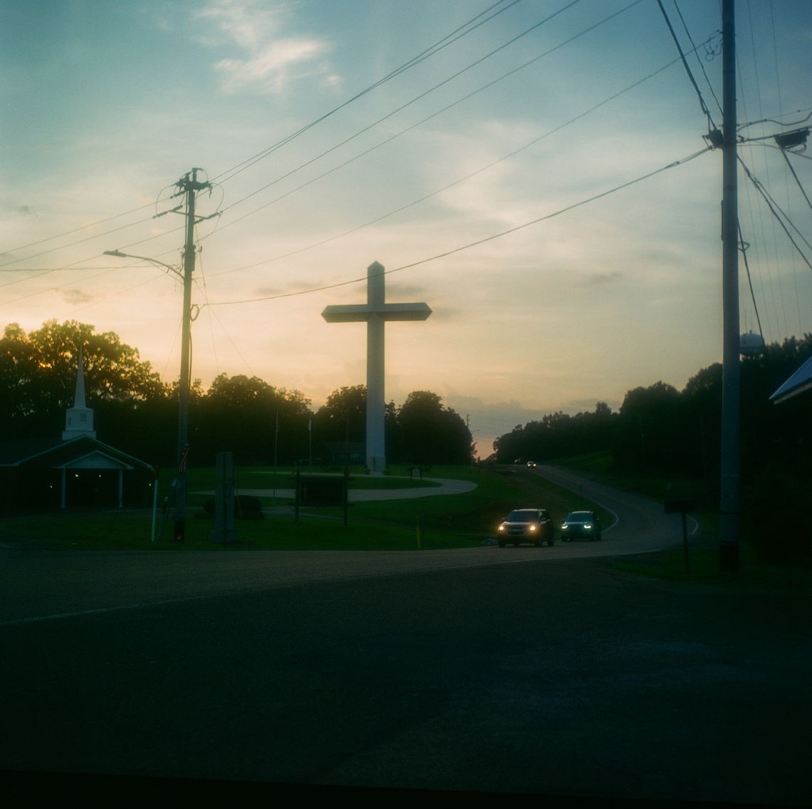 A triptych of three color photos of a large cross next to a road and a church. 
