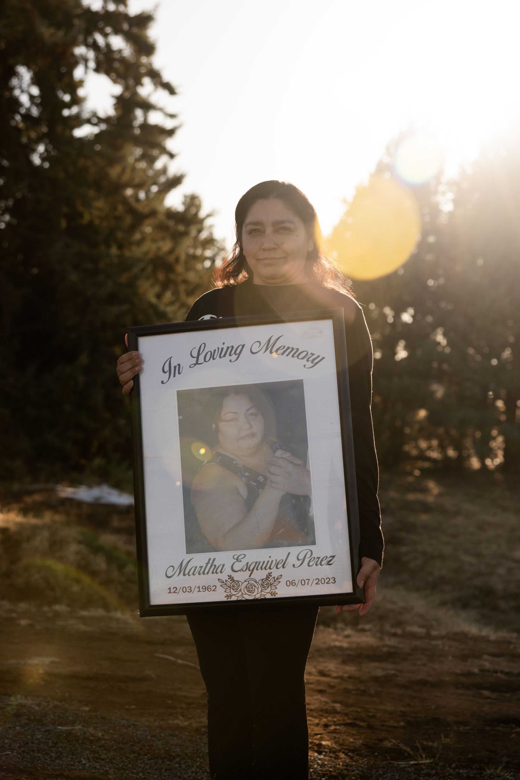 A photo shows a woman with medium-toned skin wearing black clothes holding a large framed print of a woman with the words “In Loving Memory” on the top of the print. The name Martha Esquivel Perez is at the bottom of the print.