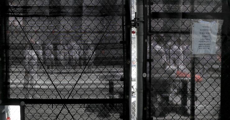 A photo shows incarcerated people seen through a fence covered in black mesh, standing in a yard outside.