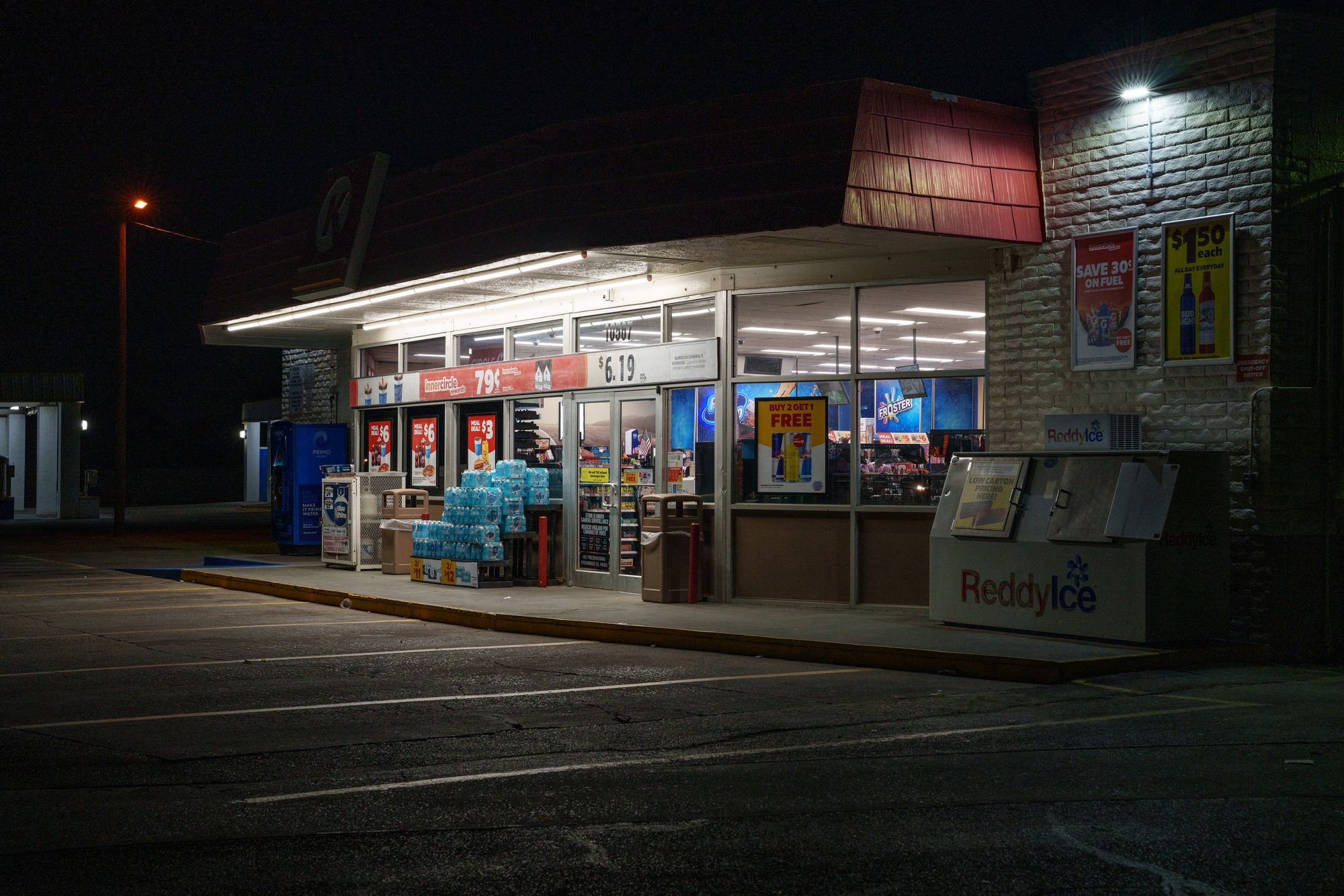 A photo shows a convenience store with a red awning, lit up at night. There is an ice merchandiser in front of it. 